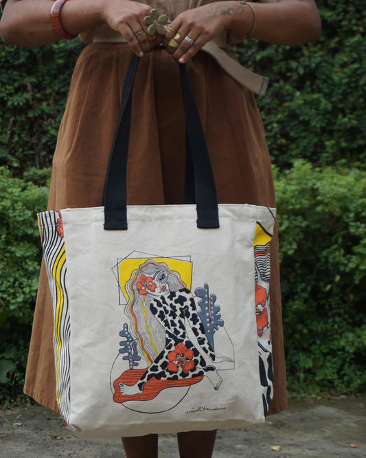 Person holding a tote bag with artistic design against a green bush background