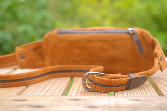 A brown suede waist bag with a matching adjustable strap and silver buckle, resting on a wooden surface with a blurred green garden background.
