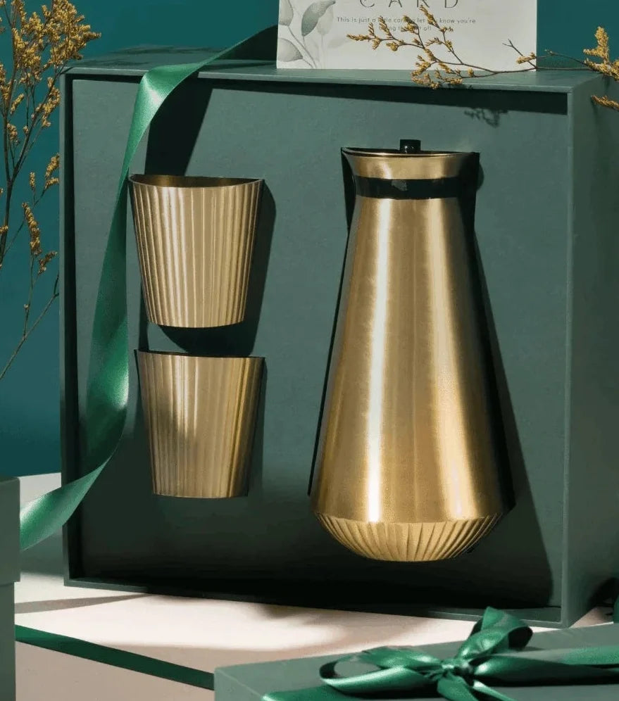 A luxury gift set featuring a metallic gold water carafe and two matching ribbed cups inside a dark green presentation box, alongside matching dark green gift boxes with green ribbons and a card that reads 'Gift Set'.