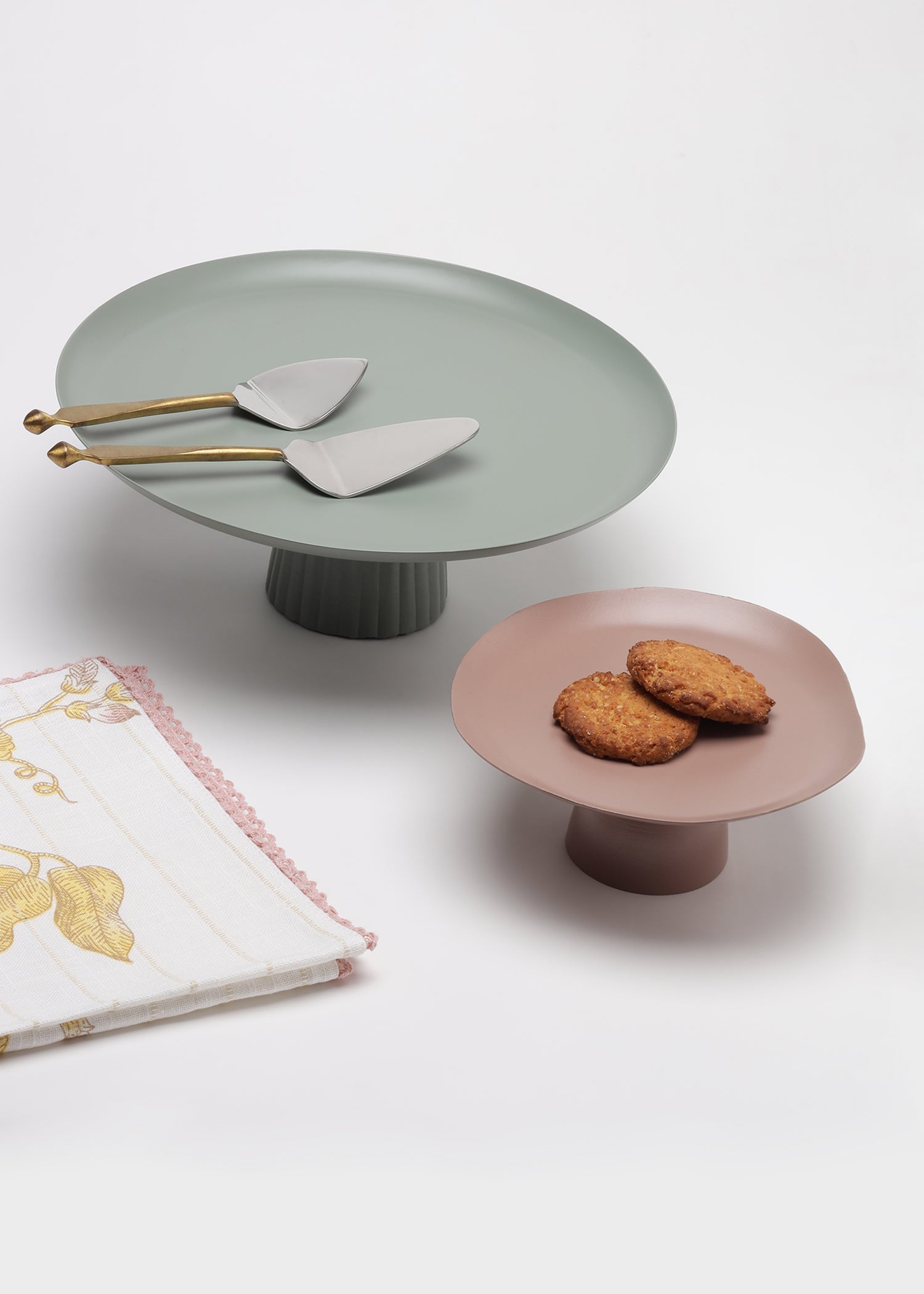 Two matte pedestal cake stands on a white studio background, one in muted sage green with gold-handled serving spoons and one in dusty pink with two cookies on top. A folded patterned napkin is partially visible at lower left.
