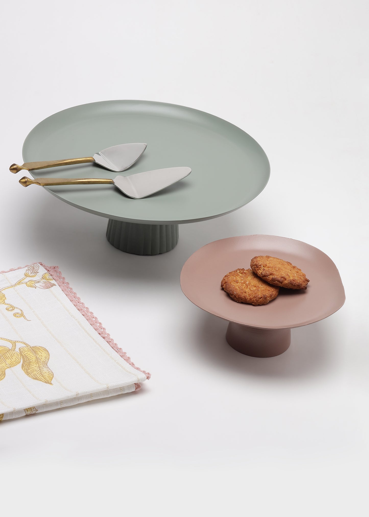Two matte pedestal cake stands on a white studio background, one in muted sage green with gold-handled serving spoons and one in dusty pink with two cookies on top. A folded patterned napkin is partially visible at lower left.