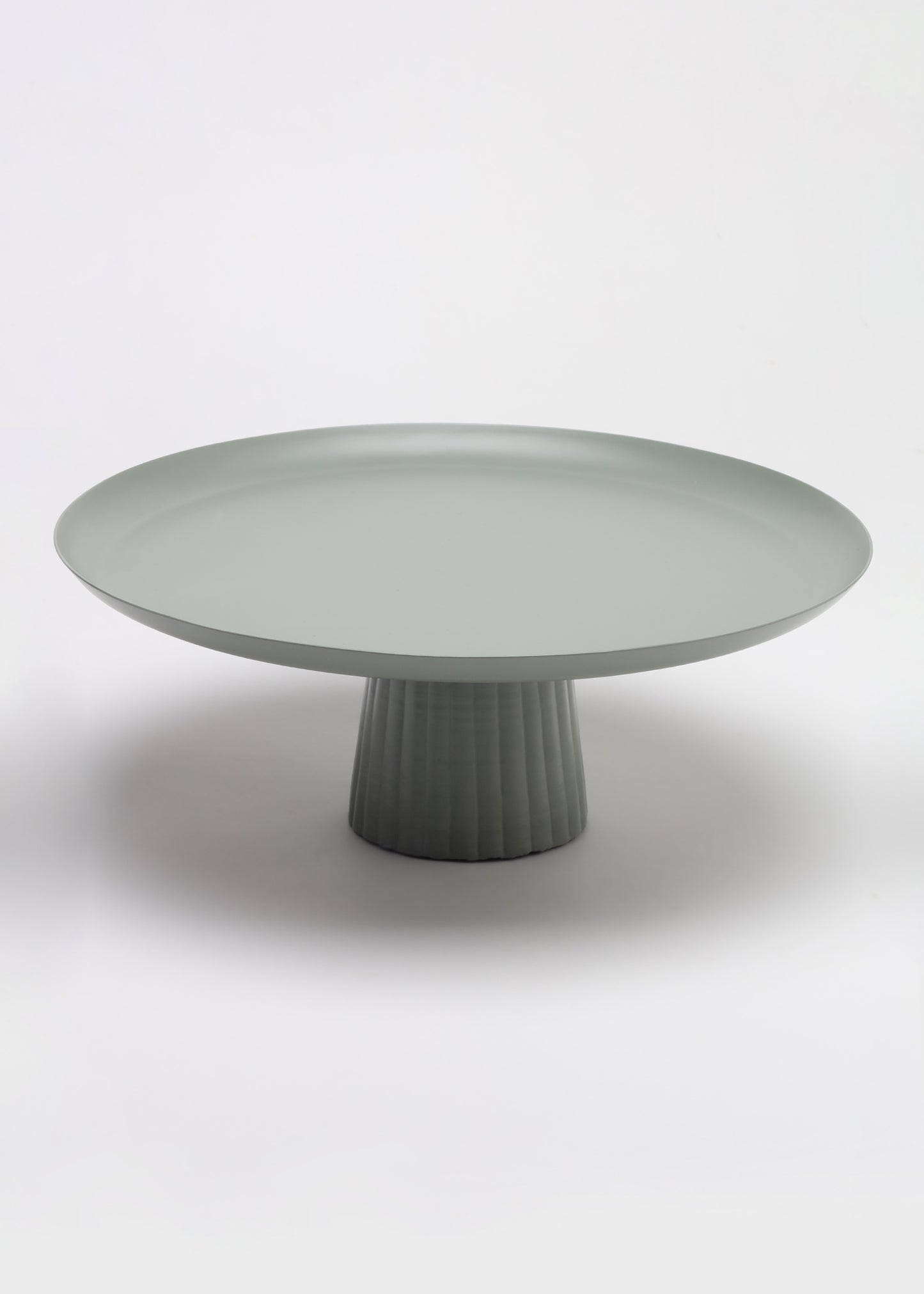 A minimalist round pedestal table in a matte sage green color, featuring a fluted cylindrical base and a wide shallow tray-like top, photographed on a seamless light gray studio background.