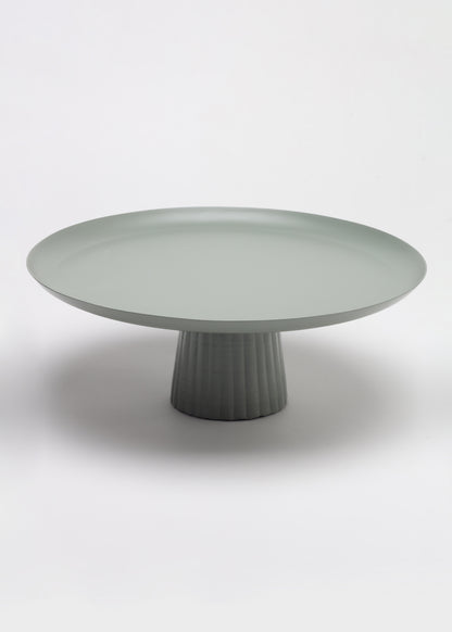 A minimalist round pedestal table in a matte sage green color, featuring a fluted cylindrical base and a wide shallow tray-like top, photographed on a seamless light gray studio background.