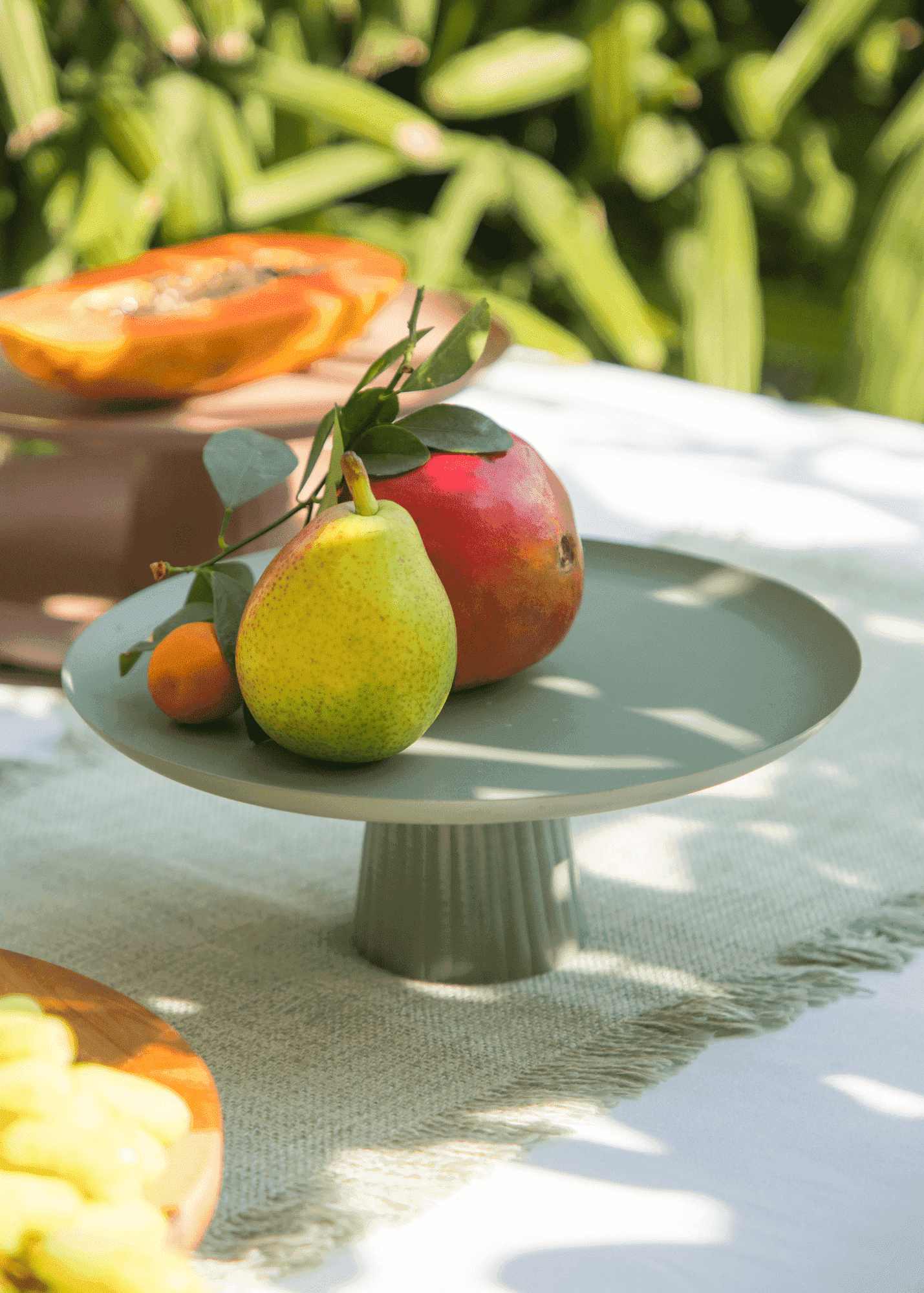 A light sage green ceramic pedestal stand holding a pear, a red mango, and a small orange, set on a white table runner with a blurred green garden background.