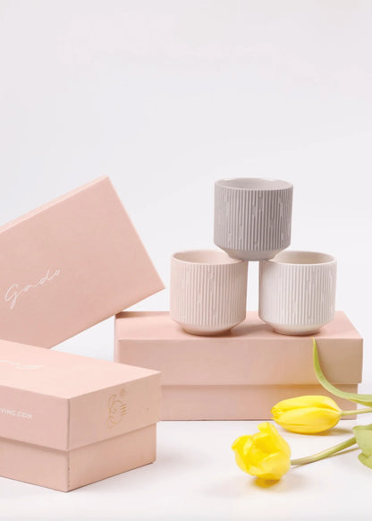 Three ribbed ceramic cups in light gray, cream, and off-white are arranged on pink gift boxes, with two yellow tulips in the foreground on a clean white background.