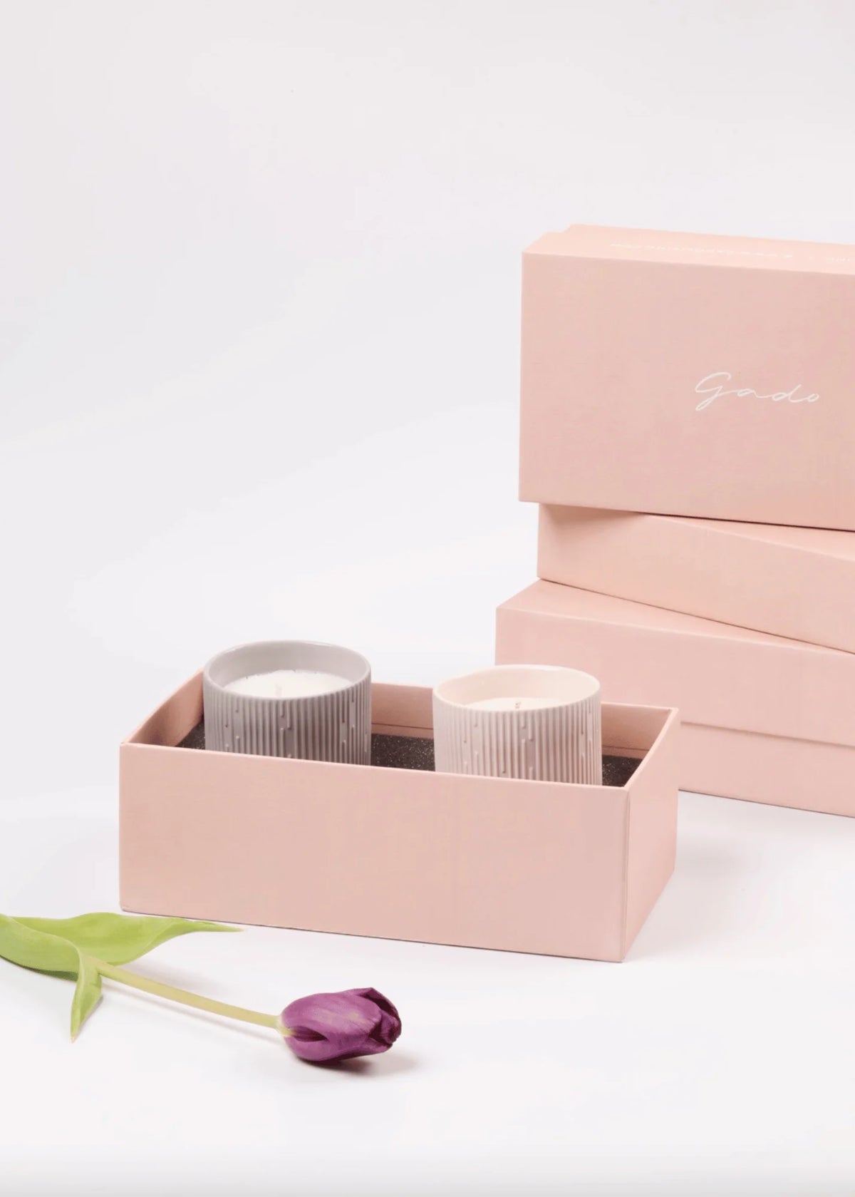 Two ribbed glass candle jars in white and light gray, displayed in a blush pink gift box with additional matching pink boxes stacked in the background and a tulip flower on the surface.