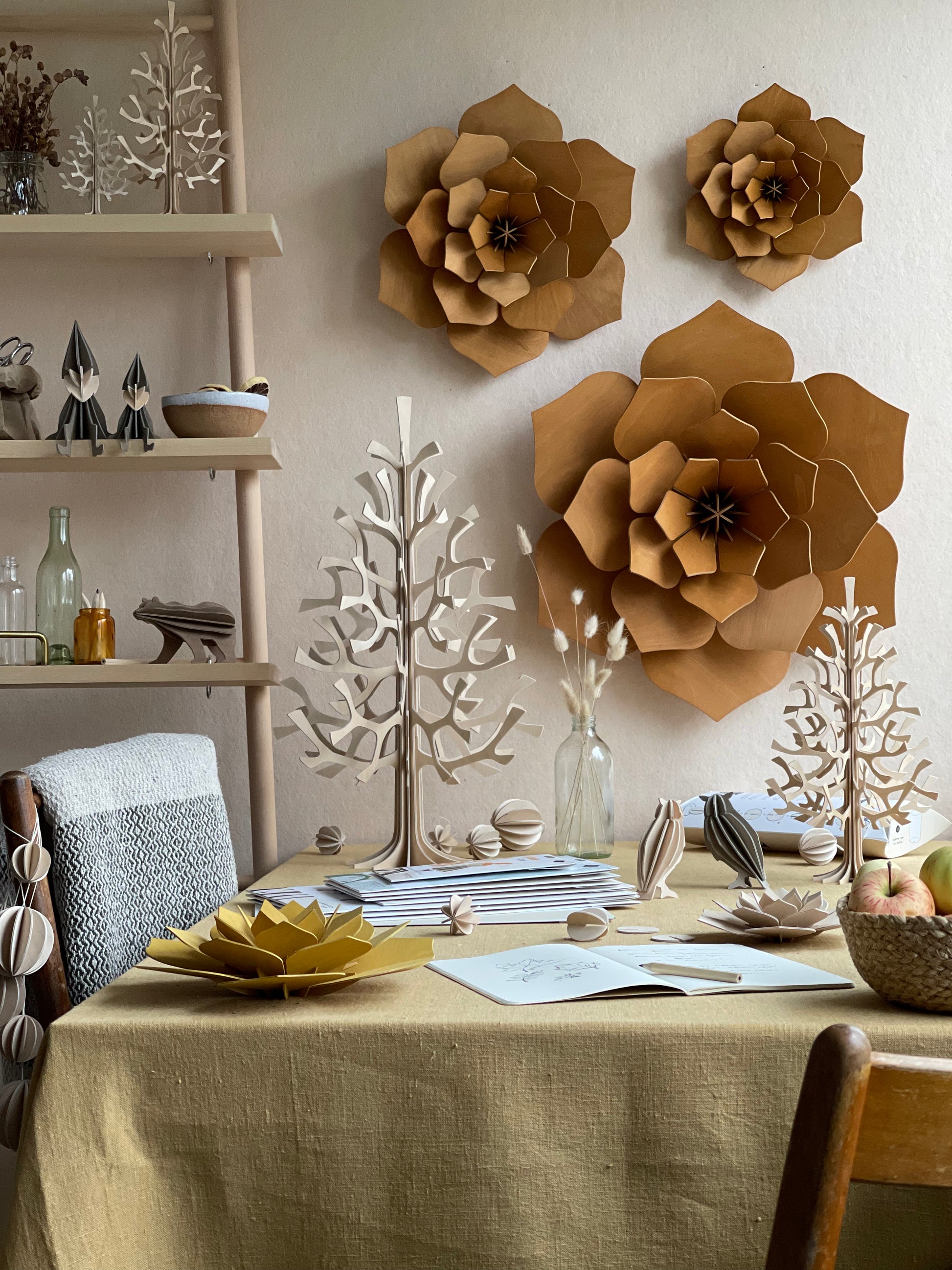 A styled dining table in a warm, rustic room decorated with large brown paper flower wall art and matching paper tree sculptures. The table is covered in a beige linen tablecloth and styled with books, small wooden objects, and a bowl of fruit.
