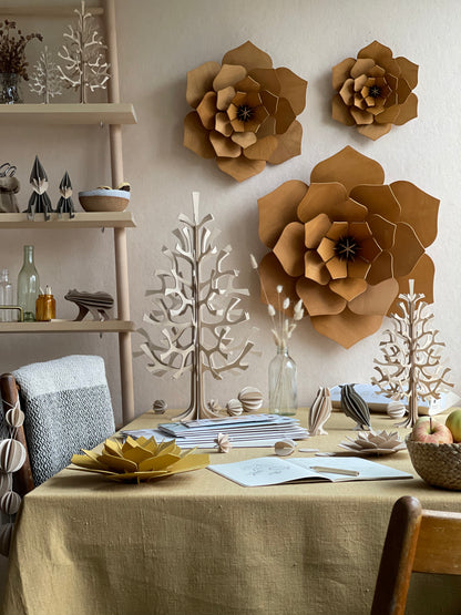 A styled dining table in a warm, rustic room decorated with large brown paper flower wall art and matching paper tree sculptures. The table is covered in a beige linen tablecloth and styled with books, small wooden objects, and a bowl of fruit.
