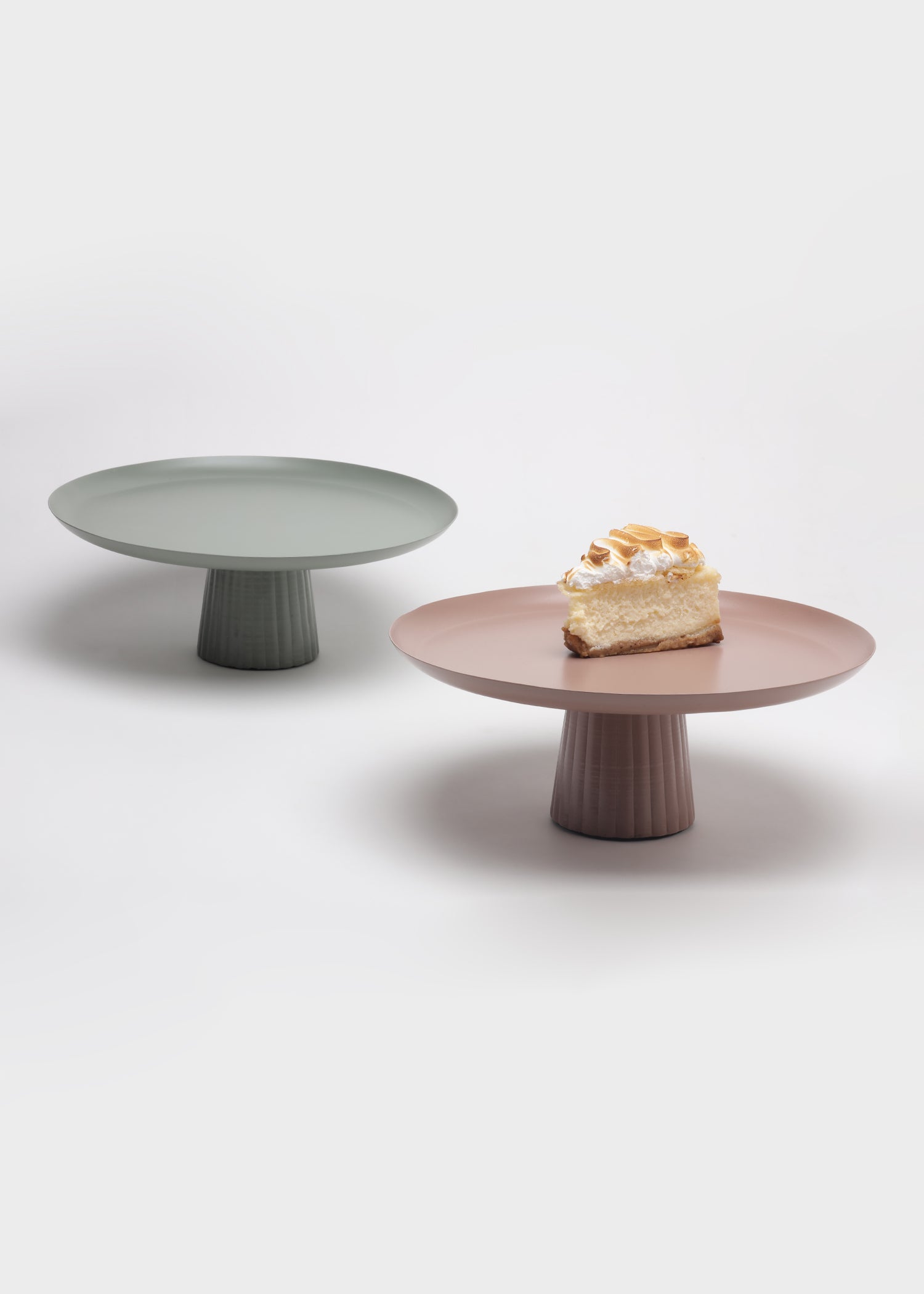 Two cake stands, one green and one brown, on a white background with a piece of cake on the brown stand.