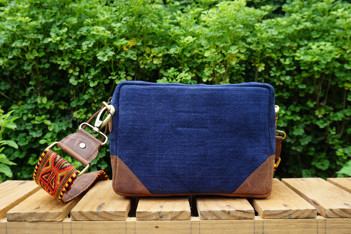 A navy blue canvas crossbody bag with brown leather corner panels and a patterned brown leather strap, resting on a wooden bench with a green leafy hedge in the background.