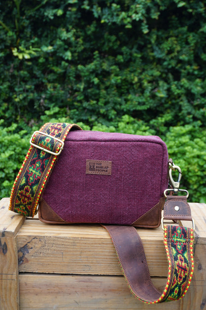 A purple woven fabric camera bag with brown leather corners and a colorful patterned webbing strap, resting on a wooden crate with a green garden background.