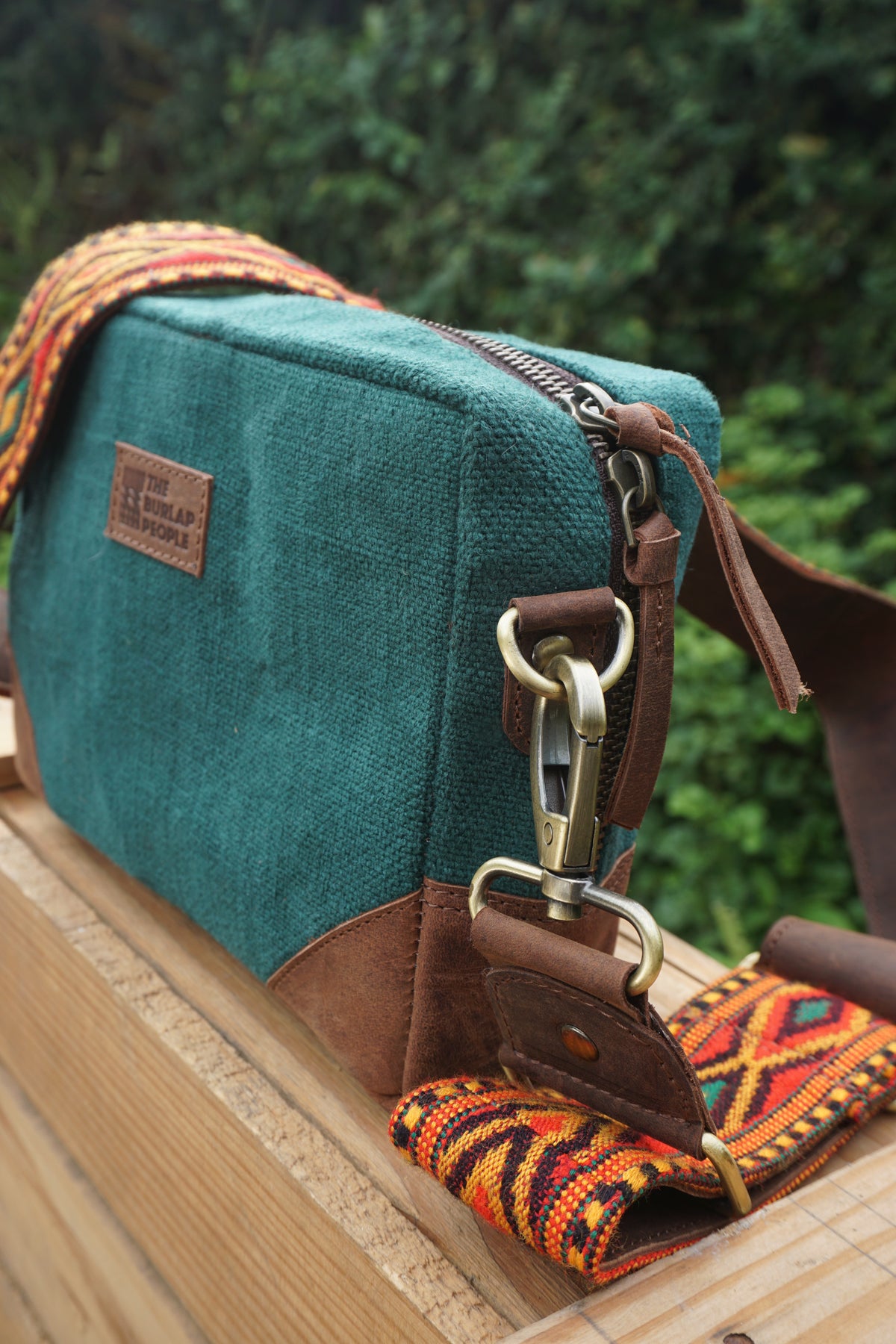 A teal canvas crossbody bag with brown leather trim and a colorful patterned woven strap, resting on a wooden surface with a blurred green outdoor background.
