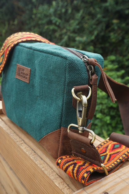 A teal canvas crossbody bag with brown leather trim and a colorful patterned woven strap, resting on a wooden surface with a blurred green outdoor background.