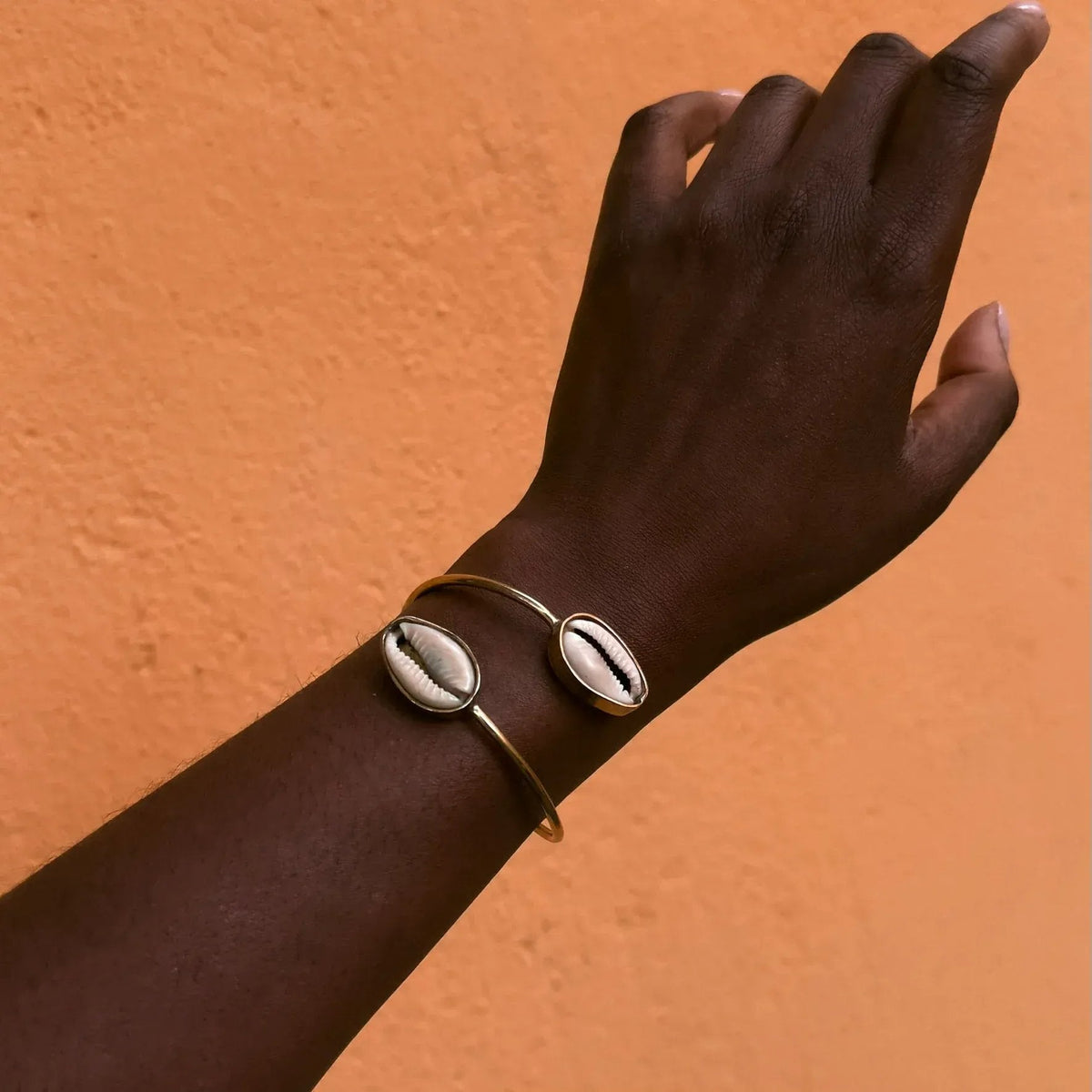 Hand wearing a gold bracelet with a cowrie shell design against an orange background