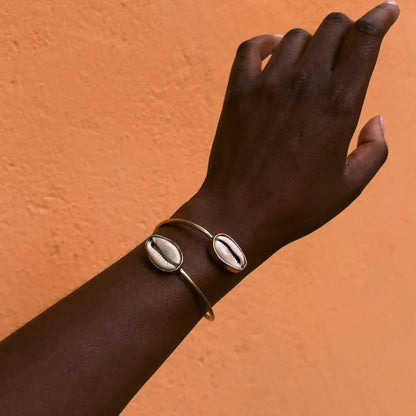 Hand wearing a gold bracelet with a cowrie shell design against an orange background