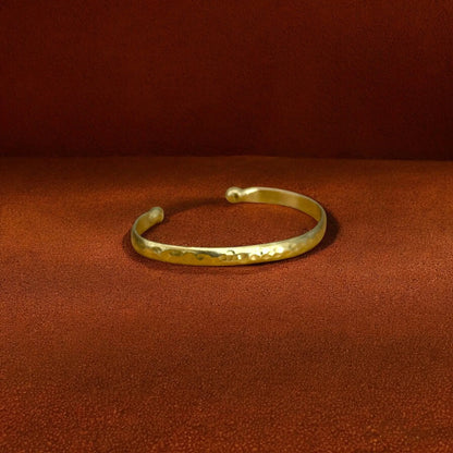 A hammered gold cuff bracelet resting on a textured brown surface against a matching brown background.