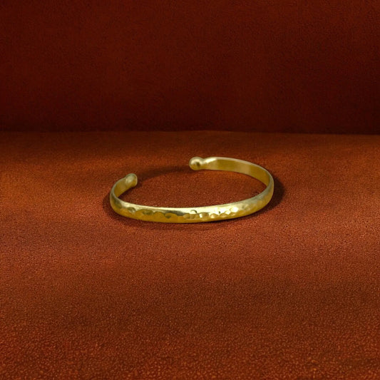 A hammered gold cuff bracelet resting on a textured brown surface against a matching brown background.
