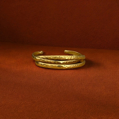 A hammered gold-tone metal cuff bracelet resting on a textured rust-brown surface.