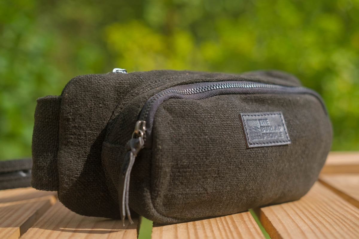 A dark olive green canvas pouch bag with a top zipper and a small rectangular brand patch, resting on a wooden surface with a blurred green outdoor background.