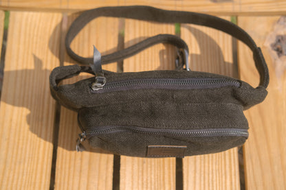 A dark olive green canvas crossbody bag with a wide adjustable strap, resting on a light-colored wooden slatted surface.