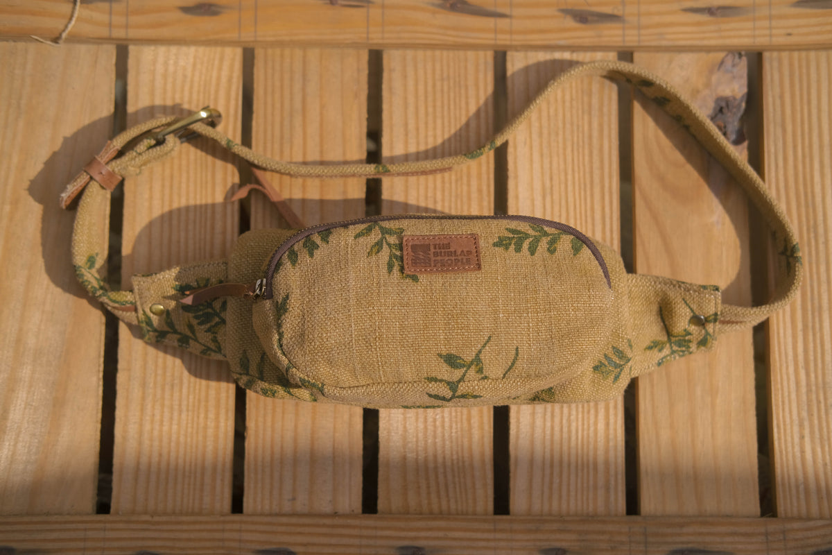 A canvas fanny pack with a green leaf pattern and a brown leather brand patch, laid flat on a wooden slatted surface.
