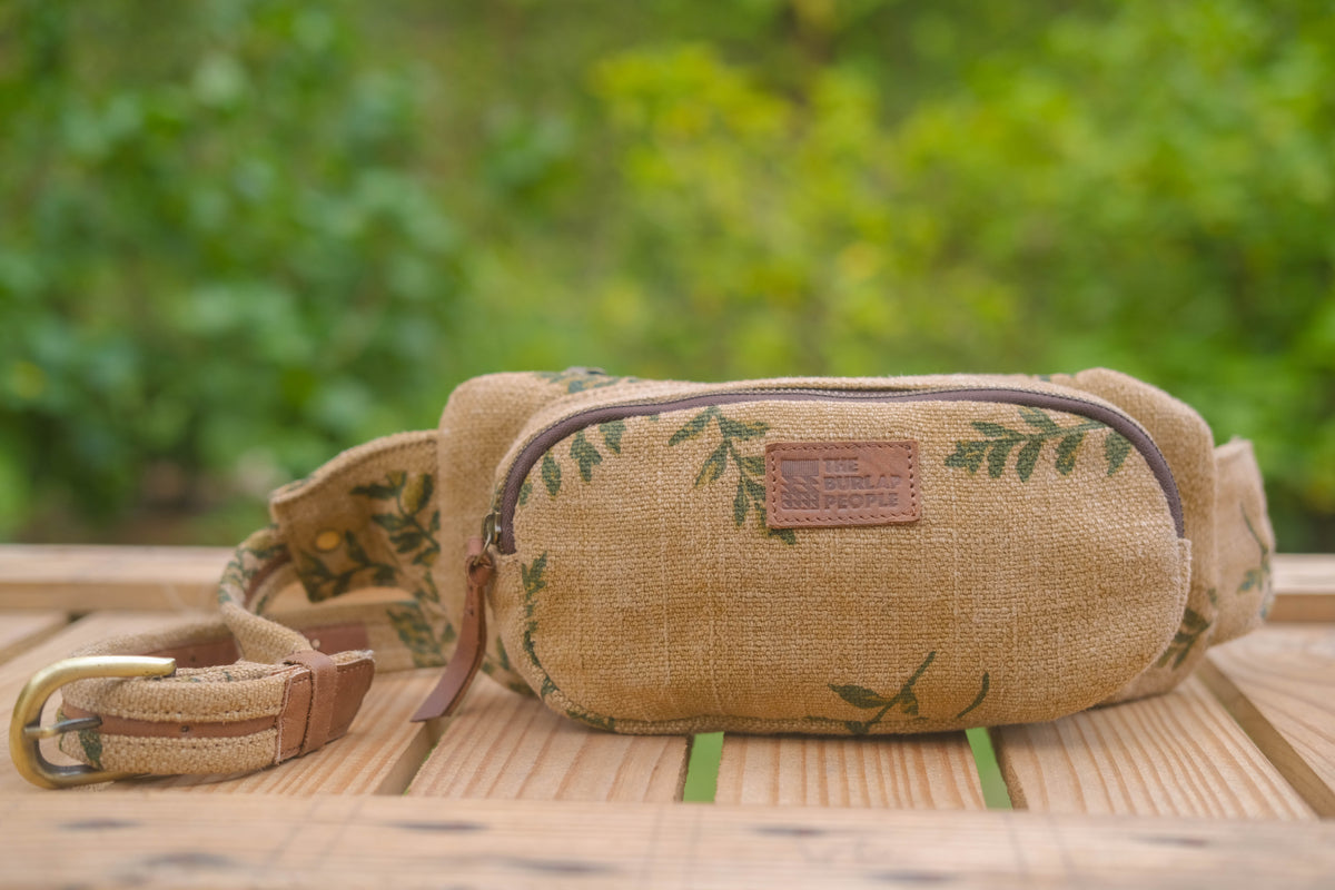 A canvas fanny pack in natural tan with green leaf patterns, featuring a brown leather brand patch that reads 'The Tree House', resting on a wooden table with a blurred green garden background.