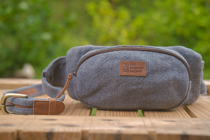 A gray canvas belt bag with brown leather trim and a brass buckle, resting on a wooden surface with a blurred green garden background. A leather patch on the front reads 'THE WAILER PEPLOE'.