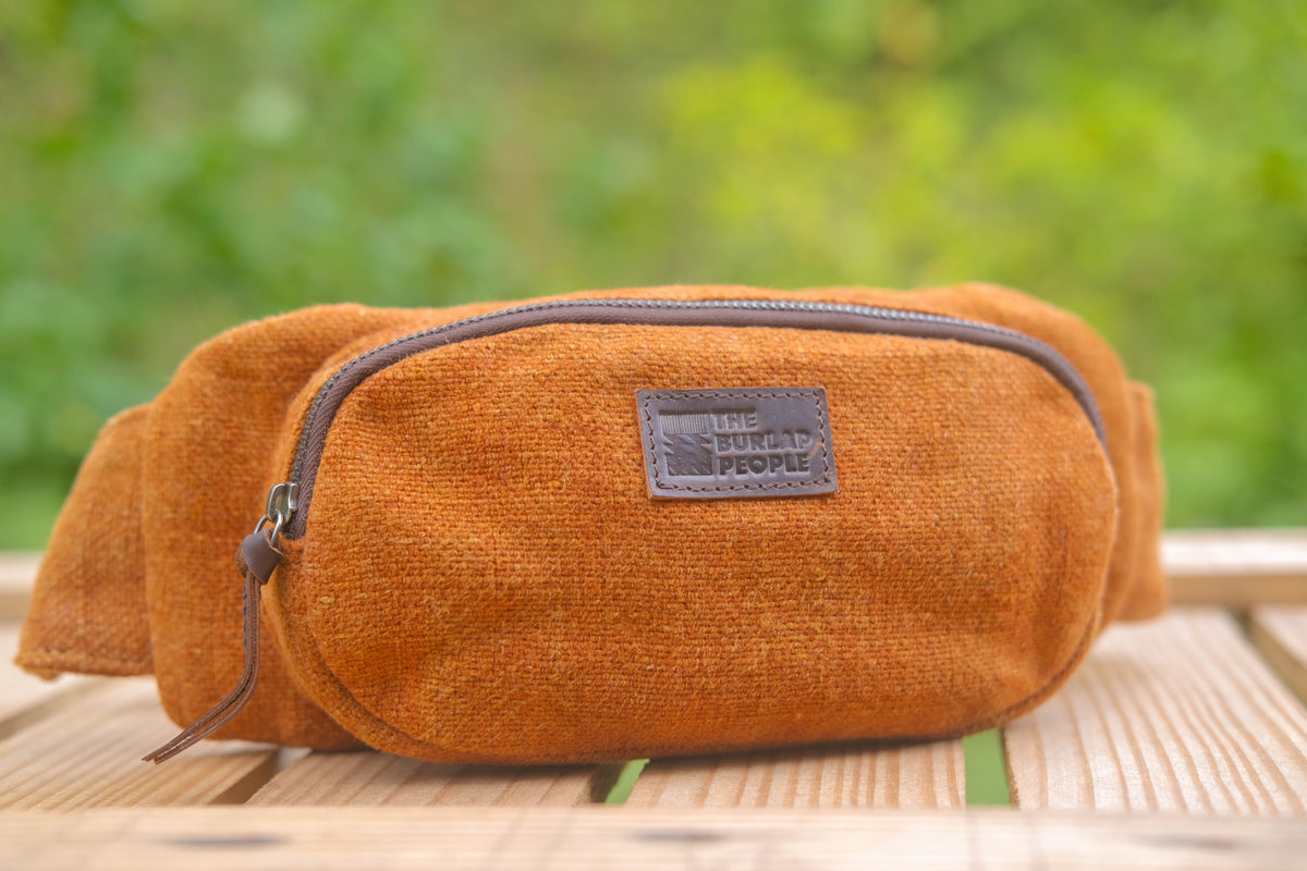An orange-brown canvas waist bag with a dark brown zipper and a brown leather patch reading 'THE BUCKET PEOPLE' on a wooden surface with a blurred green background.
