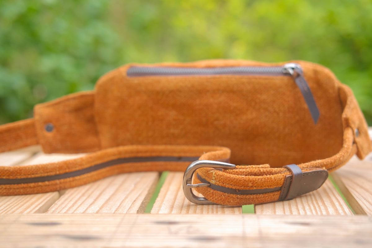 A brown suede waist bag with a matching adjustable strap and silver buckle, resting on a wooden surface with a blurred green garden background.