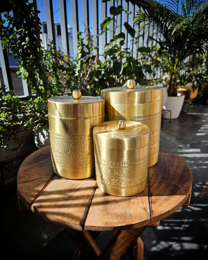 Three brass storage containers with engraved patterns and small wooden lids, displayed on a round wooden table in a sunlit patio setting with green plants in the background.