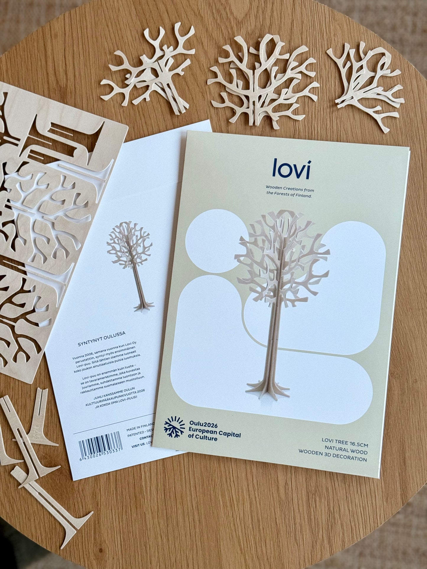 A flat lay of a wooden decorative kit on a round wooden table, showing a printed instruction booklet titled 'lovi', laser-cut wooden tree and coral shapes, and wooden dowels.
