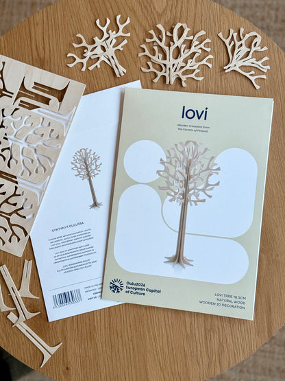 A flat lay of a wooden decorative kit on a round wooden table, showing a printed instruction booklet titled 'lovi', laser-cut wooden tree and coral shapes, and wooden dowels.