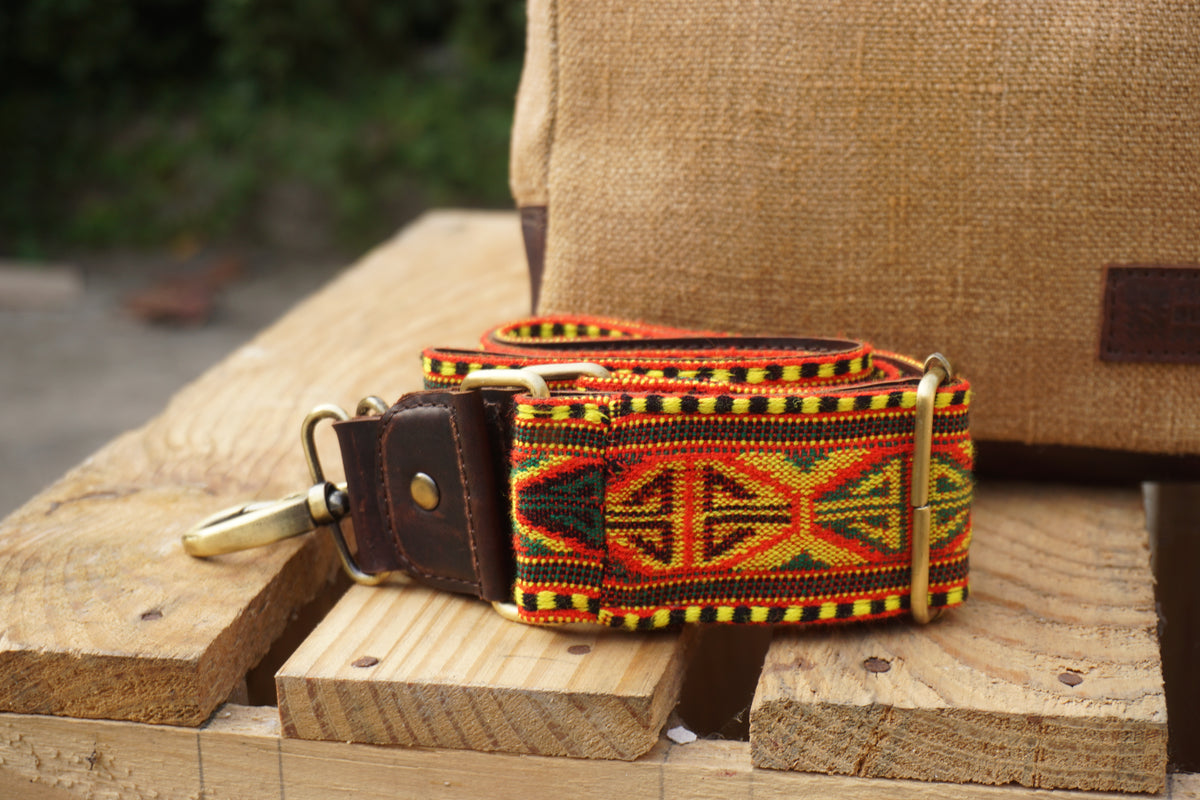 A woven belt with a Southwestern geometric pattern in red, yellow, green, and black, featuring a dark brown leather strap and brass hardware, resting on wooden blocks.