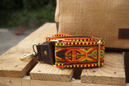A woven belt with a Southwestern geometric pattern in red, yellow, green, and black, featuring a dark brown leather strap and brass hardware, resting on wooden blocks.