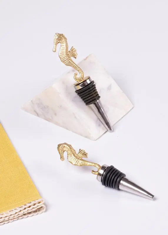 Two gold-toned seahorse-shaped wine bottle stoppers with black rubber gaskets and silver metal points, displayed on a white marble tile and a white surface with a gold textured prop partially visible at the left edge.
