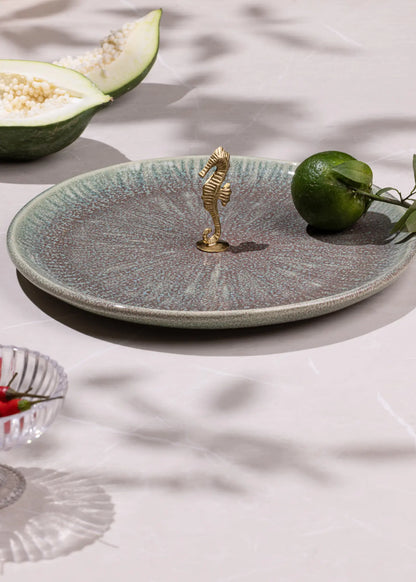 Decorative ceramic plate with a gold seahorse charm on a light surface with fruits.