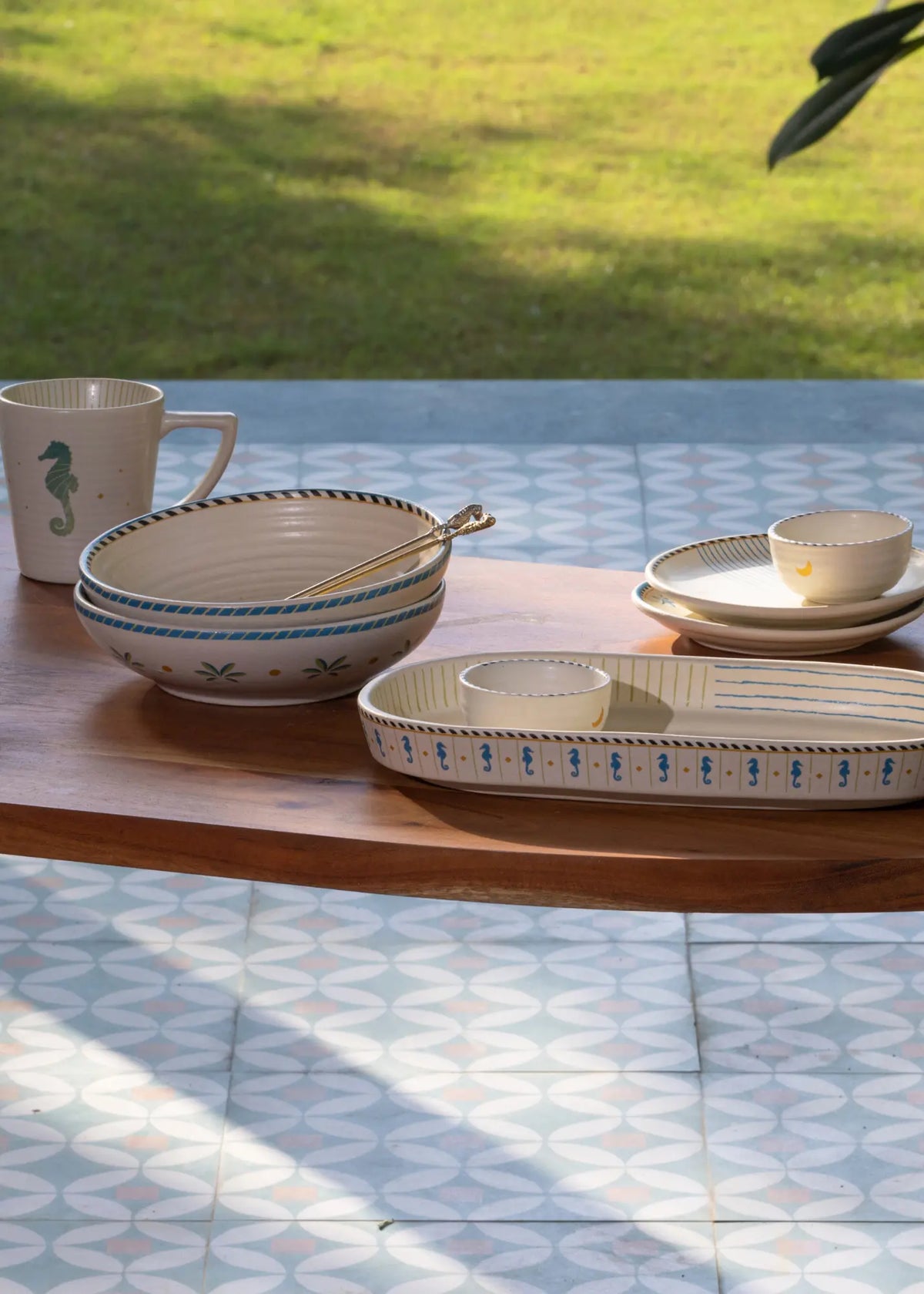 A set of beige ceramic tableware with blue rope and floral patterns, including a mug, bowls, plates, and a serving tray, arranged on a wooden table outdoors with a grassy lawn in the background.