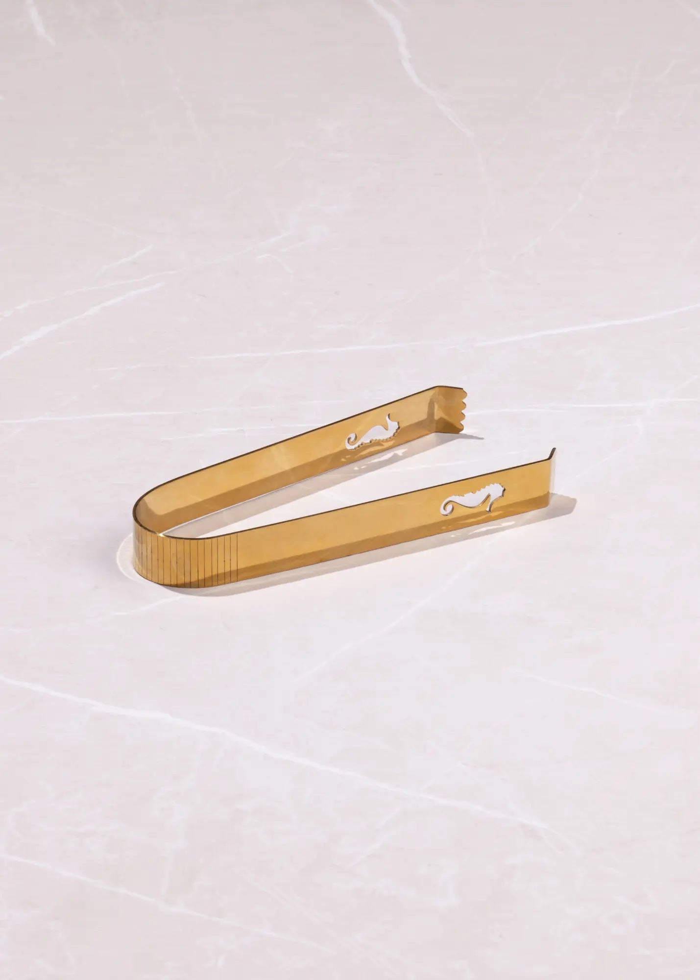 Gold metal cocktail strainer tongs with a textured ribbed handle and small cutout details, resting on a light beige marble surface.