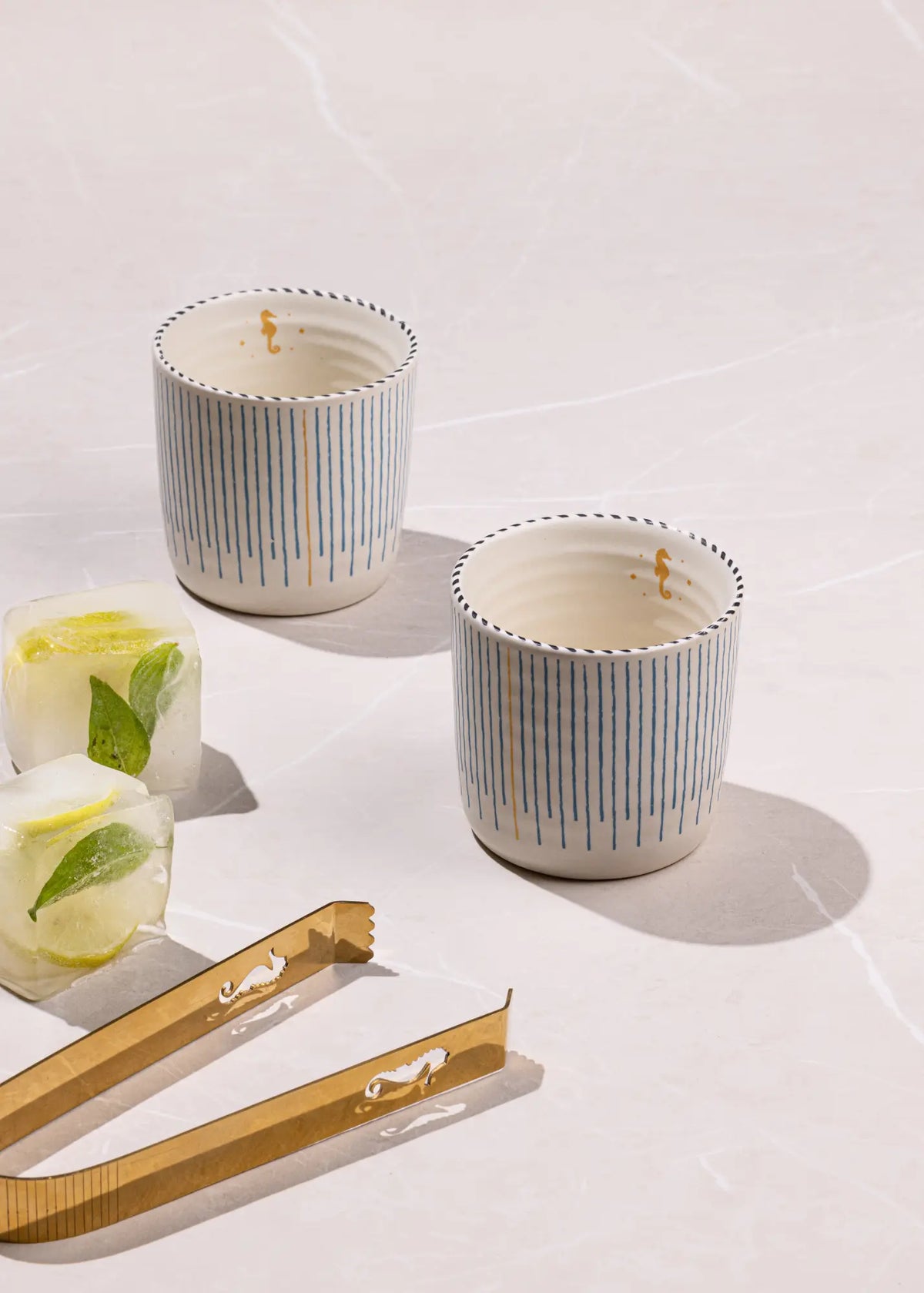 Two small white ceramic tumblers with vertical blue stripes and gold rim detailing, styled on a light marble surface with gold-toned tongs and ice cubes with mint leaves.