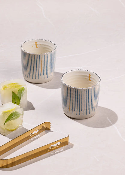 Two small white ceramic tumblers with vertical blue stripes and gold rim detailing, styled on a light marble surface with gold-toned tongs and ice cubes with mint leaves.
