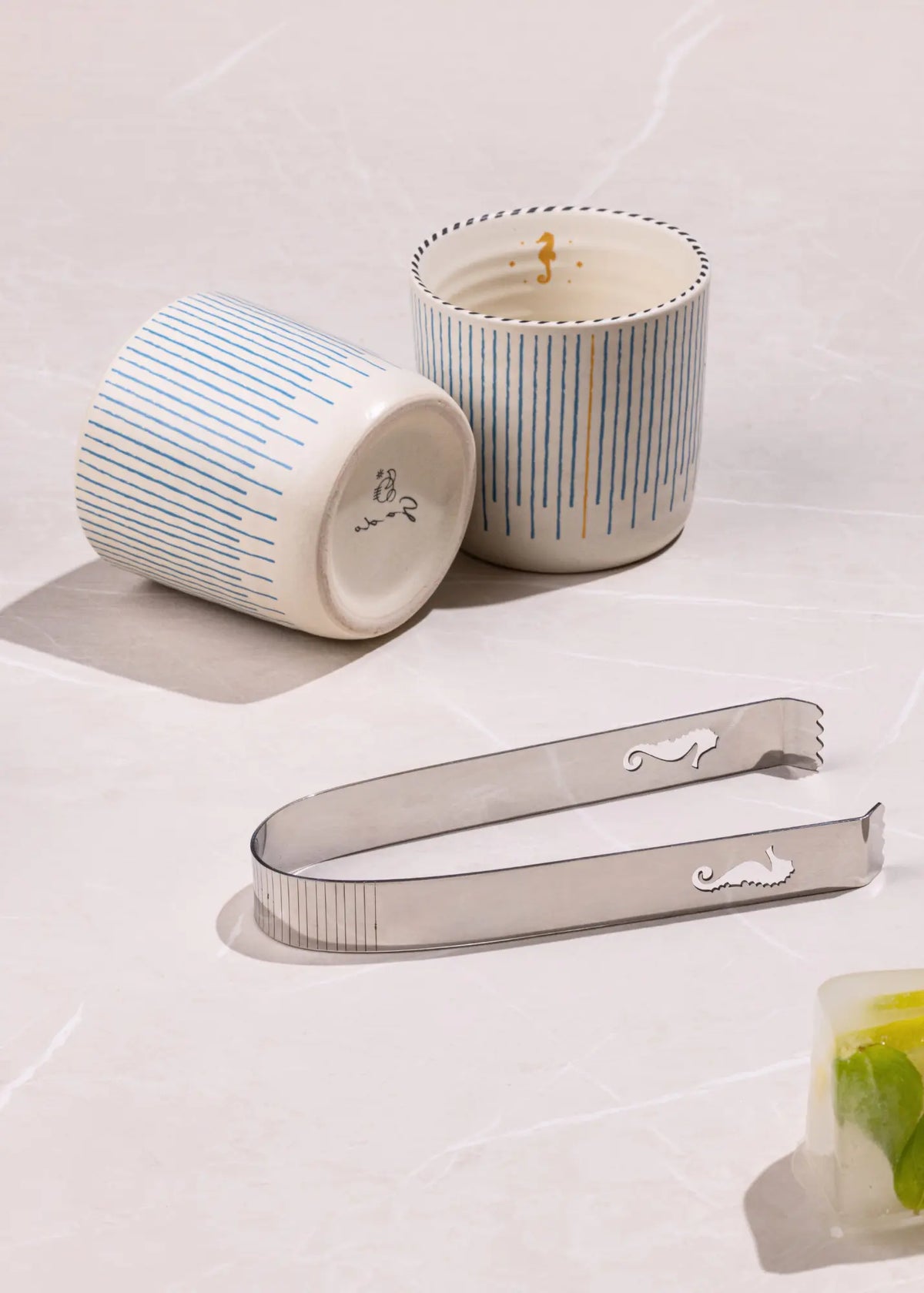 Two striped ceramic candles and a stainless steel cocktail strainer on a light marble surface.