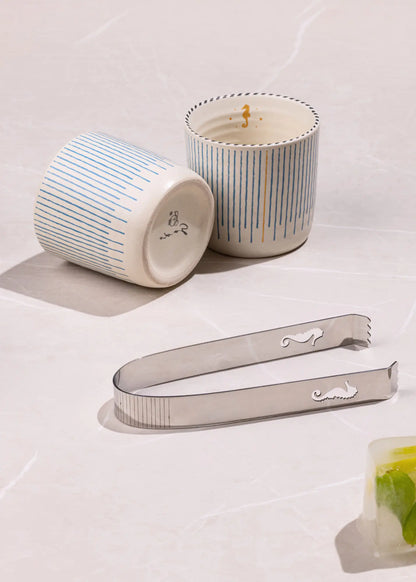 Two striped ceramic candles and a stainless steel cocktail strainer on a light marble surface.