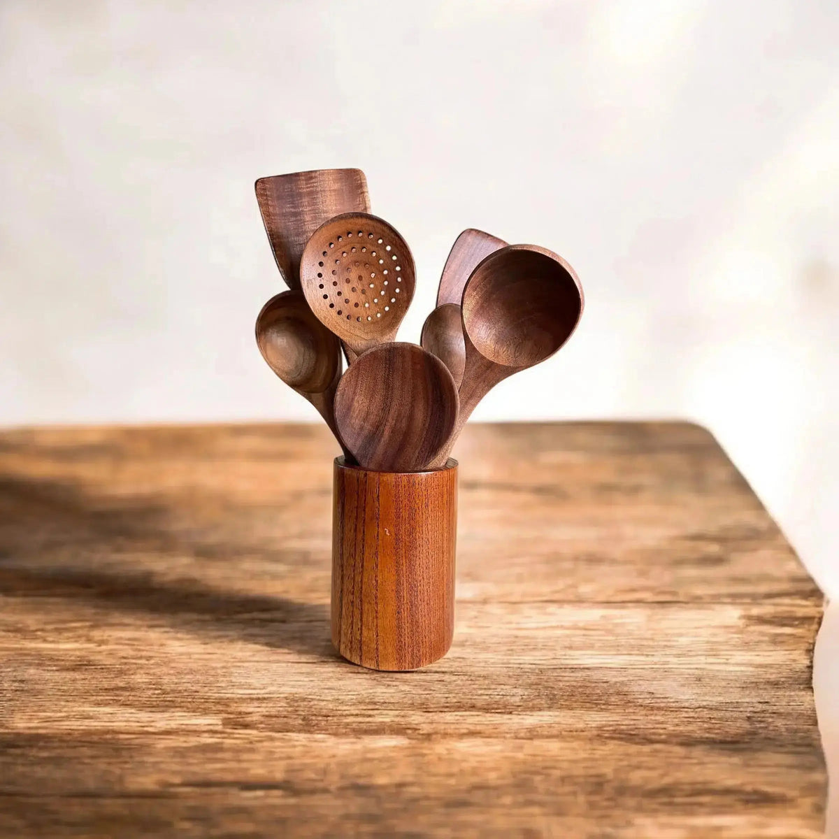A set of wooden kitchen utensils, including spoons and a strainer, arranged in a small wooden holder on a rustic wooden table against a soft, light background.