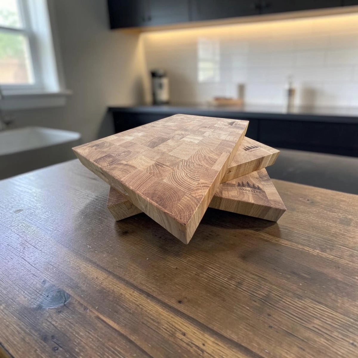 Three stacked rectangular wooden cutting boards with visible wood grain patterns, displayed on a dark wood kitchen table with a blurred kitchen background.