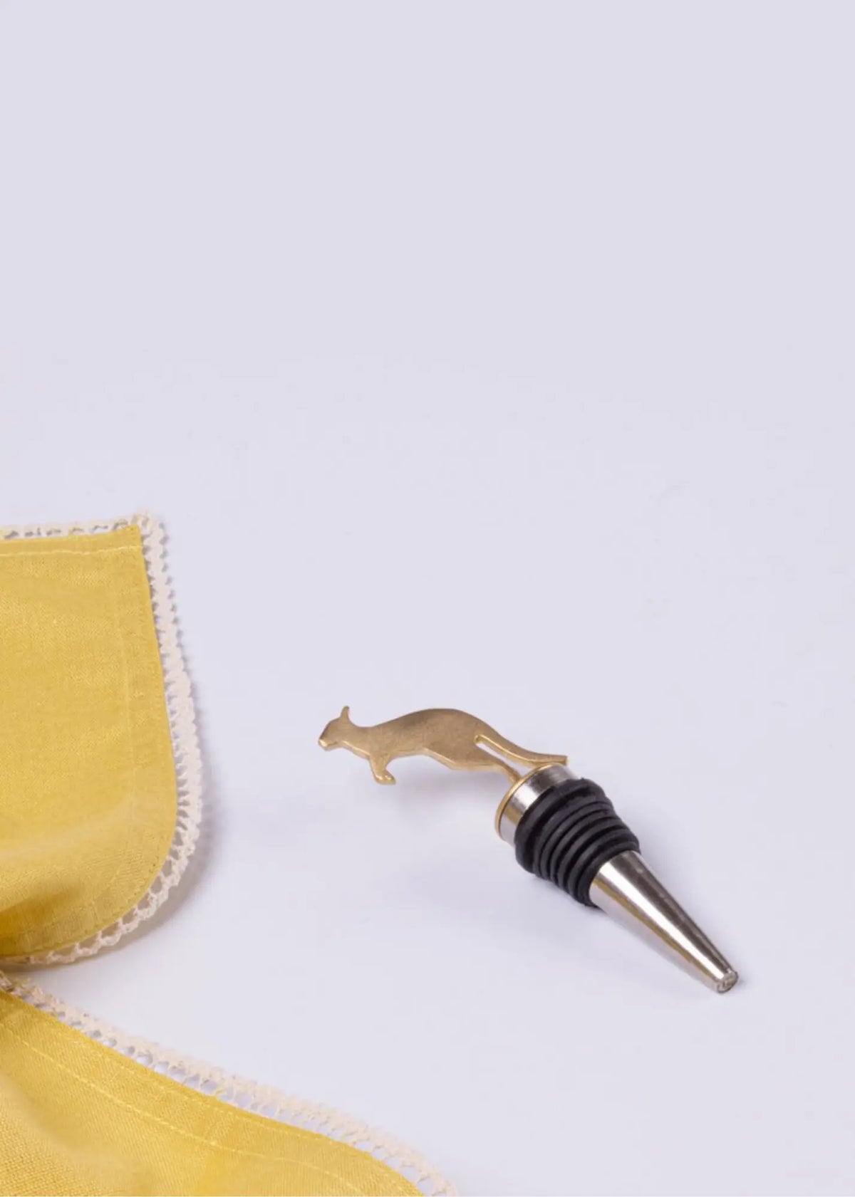 A brass bottle stopper shaped like a kangaroo with a black rubber gasket and silver metal base, shown on a light gray background next to a yellow textured fabric corner.