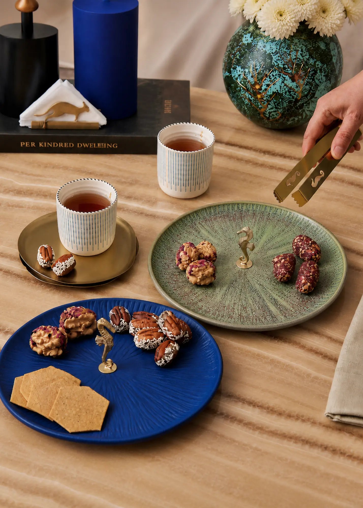 Three round ceramic plates in blue, green, and white are arranged on a wooden table with tea cups, nuts, and crackers. A hand holding a gold-toned utensil enters from the right side.