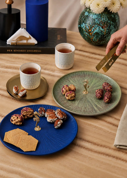 Three round ceramic plates in blue, green, and white are arranged on a wooden table with tea cups, nuts, and crackers. A hand holding a gold-toned utensil enters from the right side.