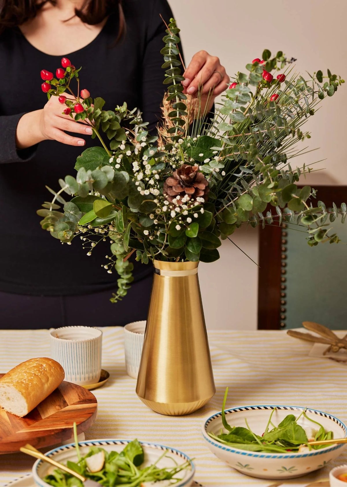 Person arranging flowers in a gold vase on a table with a bowl of salad and bread.