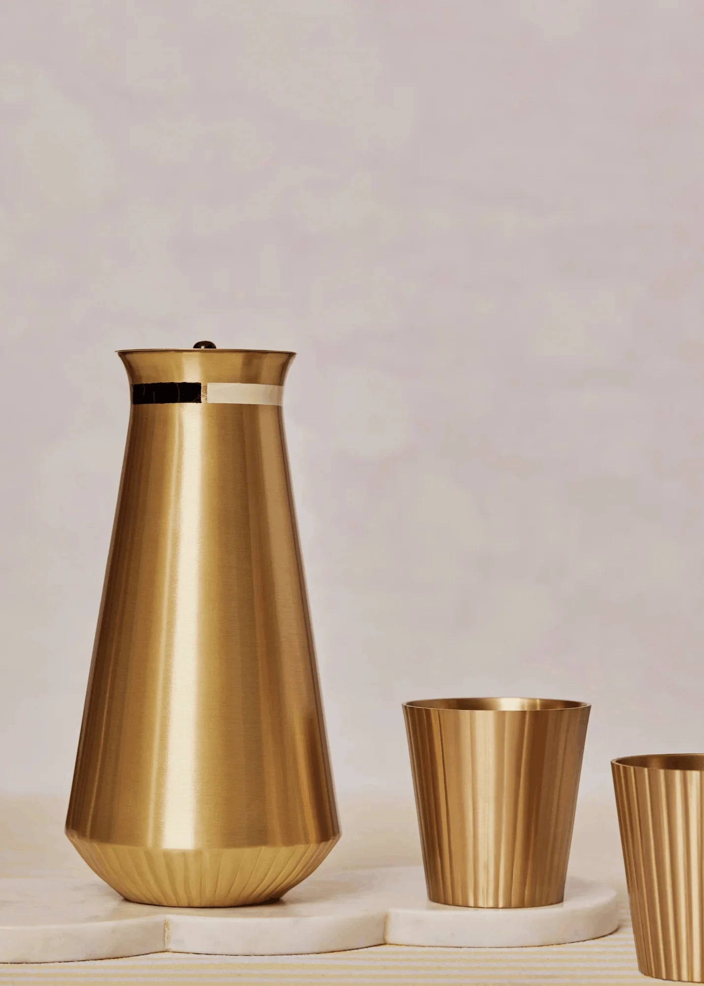 A brushed gold metal carafe with a matching lid sits on a white stone slab next to two matching fluted gold cups against a soft beige background.