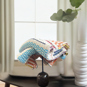 A multicolored geometric patterned Sujni quilt displayed on a stand in a room, with a window and plant in the background.